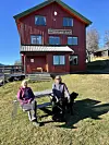 To personar sit på ein benk framfor ein raud skulebygning med ein svart hund.