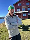 Mann med grøn lue står framfor ein raud skulebygning ved vatnet.