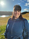 Kvinne i blå dongerikjole står ved eit vatn i solskin.