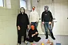 Fire maskerte personar poserer i eit flislagt rom, med fargerike gjenstandar på golvet.