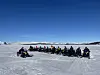 Gruppe på snøskuterar står samla på eit islagt vatn framfor snødekte fjell.